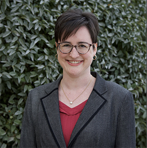 Carolyn, a woman with light skin, a dark pixie cut, and glasses. She wears a bright pink top under a trim gray blazer.